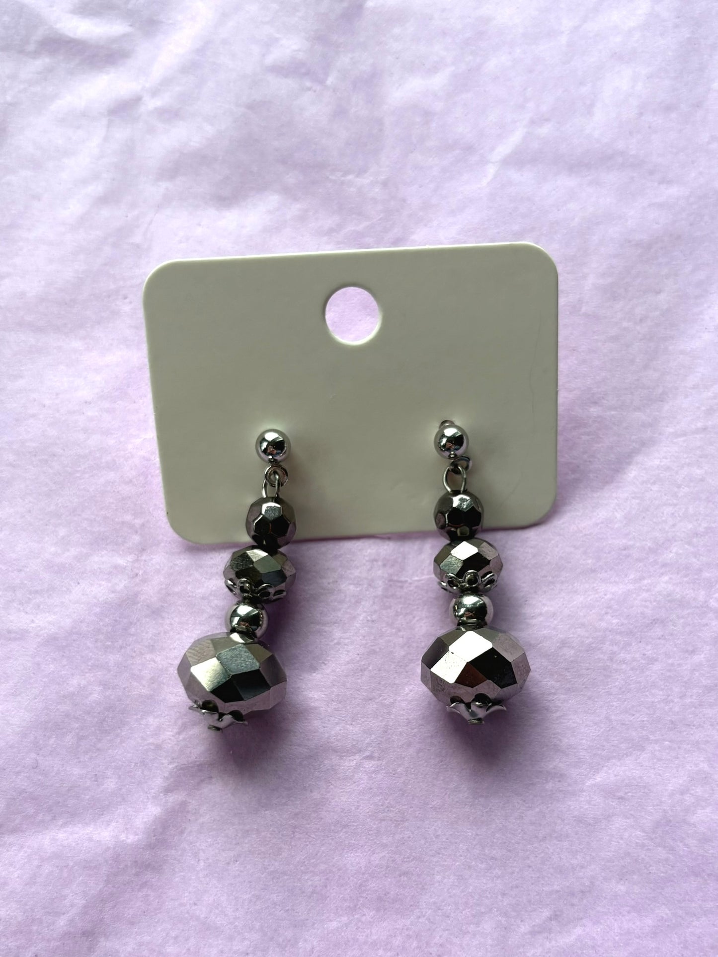 Pair of silver beaded earrings on a white card against a light purple background